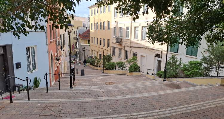 Gepflasterte Treppe, gesäumt von pastellfarbenen Gebäuden und belaubten Bäumen in einer mediterranen Altstadt.