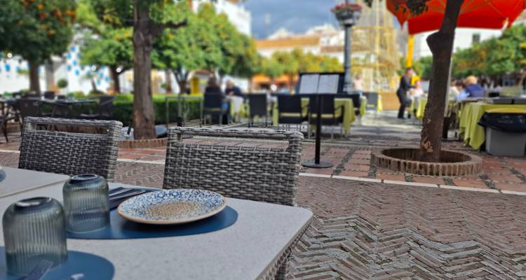 Tischgedeck im Freiluftrestaurant mit geflochtenen Stühlen und unscharfen Gästen auf einem sonnigen Platz.
