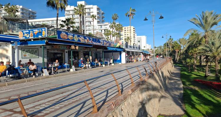Von Palmen gesäumte Strandpromenade mit Freiluftcafés und strahlend blauem Himmel.