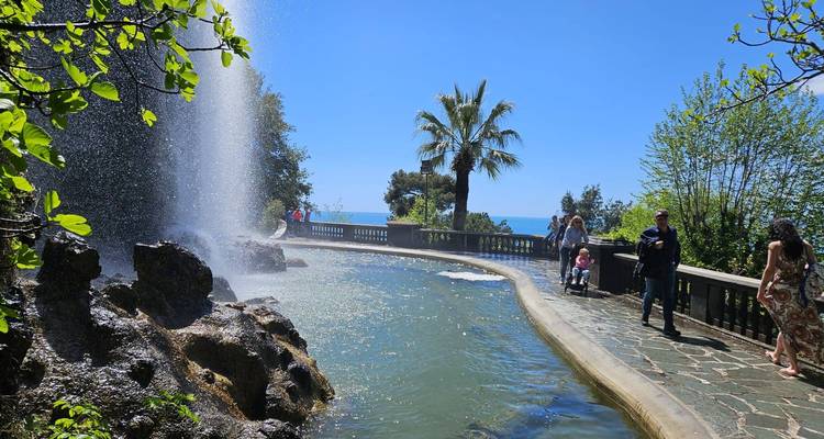 Park fountain spraying mist beside a palm tree promenade with families and sea visible beyond.