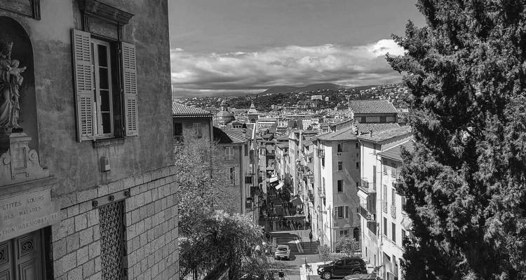 Black-and-white cityscape overlooking a tiled roof neighborhood toward distant hills under dramatic clouds.