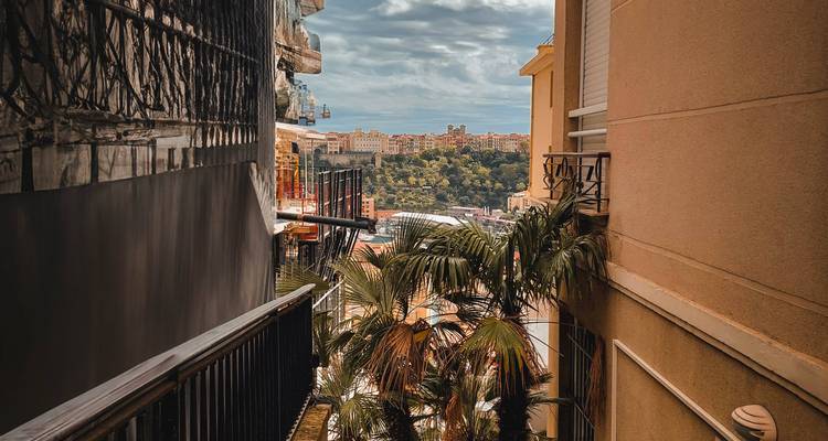 View down a narrow gap between buildings toward palm trees and a distant hillside cityscape under dramatic sky.