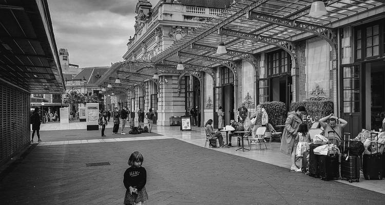 Monochrome scene of a historic railway station forecourt with travellers, luggage and a small child in foreground.
