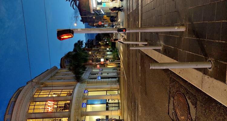 Evening street corner in Nice with H&M store, palm trees and a red pedestrian light glowing.