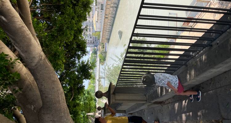 Un enfant assis sur un banc au bord de la rivière sous un grand arbre tandis qu'un adulte pointe du doigt vers l'eau