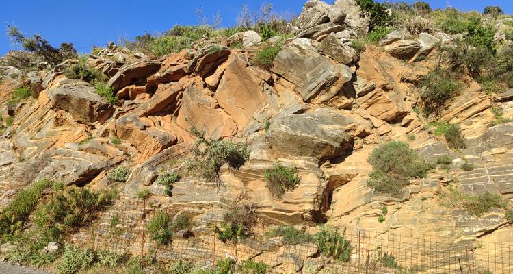 Sunny roadside cutting revealing layered orange and grey rock formations behind a mesh fence.