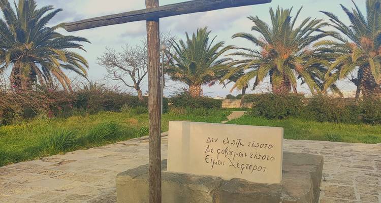 Stone plinth with Greek inscription and wooden cross surrounded by palms and greenery at sunset light.