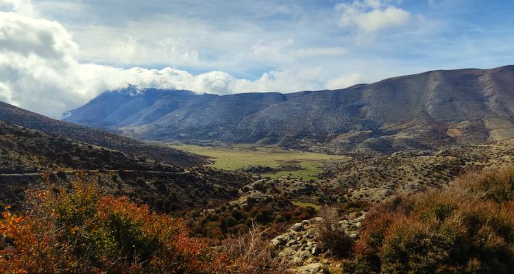 Wide mountain valley carpeted in green pastures, framed by autumn shrubs and cloud-topped ridges.