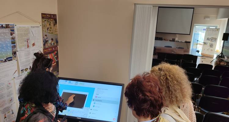 Four women watch a touchscreen presentation in a small conference room with chairs and posters.