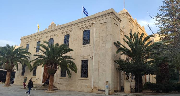 Elegant cream-stone public building with Greek flag and palm trees, a passer-by in front.