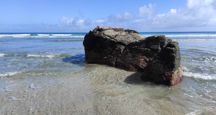 Rocher sombre isolé se dressant dans une eau de mer peu profonde et cristalline avec des vagues douces et un horizon bleu.