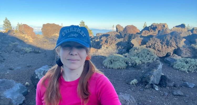 Selfie d'une femme portant un haut rose et une casquette debout sur un terrain volcanique foncé avec des arbustes dispersés.