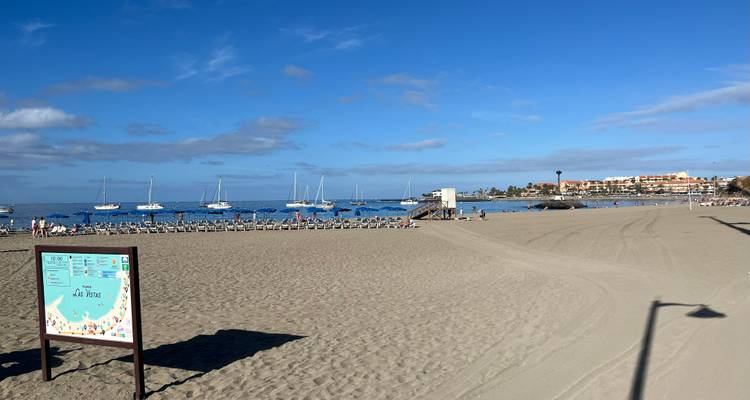Plage de sable fin avec des rangées de transats, des voiliers amarrés et une ville balnéaire au loin.