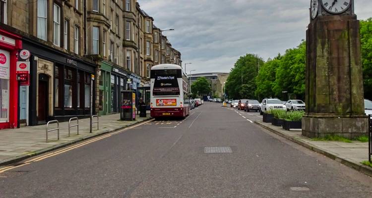 Urban street with double-deck bus, stone shopfronts, and cloudy skies.