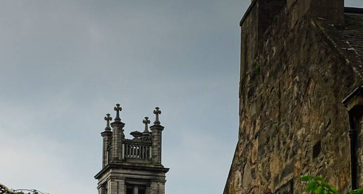 Stone bell tower with ornate crosses rising above a weathered stone building under grey sky.