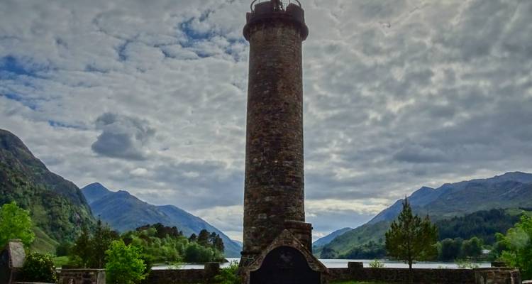 Tall stone monument column silhouetted against dramatic mountain and loch backdrop.