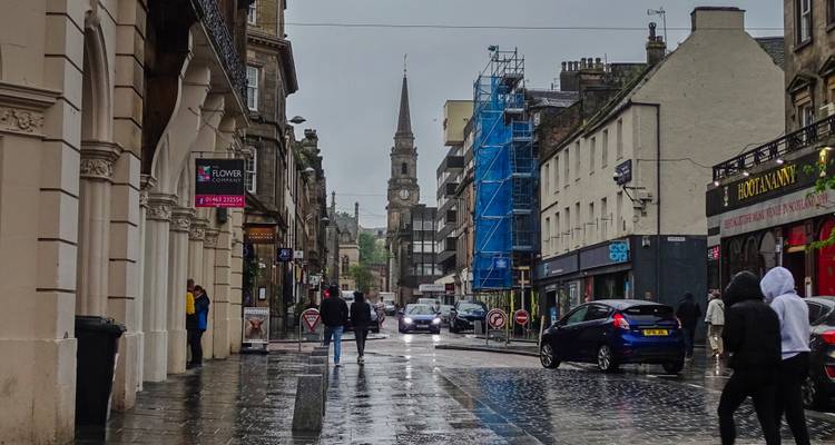 Rain-soaked city street with shoppers, cars and a distant church spire.