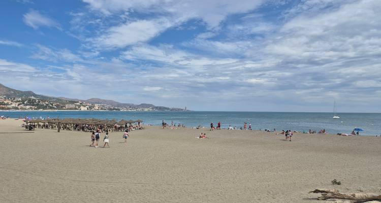 Wide sandy beach with sunbathers, swimmers, and umbrellas under partly cloudy sky