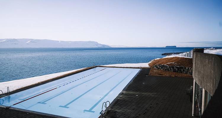 Piscine à débordement en bord de mer surplombant un fjord tranquille et des falaises enneigées lointaines sous un ciel dégagé.