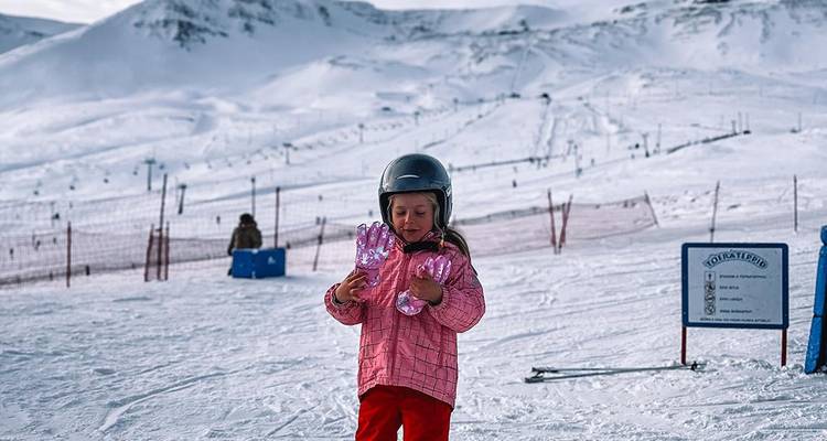 Jeune enfant en casque et combinaison de ski rose se tient sur une piste de ski avec des jouets à la main.