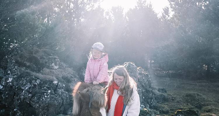 La mère guide un petit cheval islandais tandis que son enfant sourit en selle au milieu des arbres baignés de soleil.