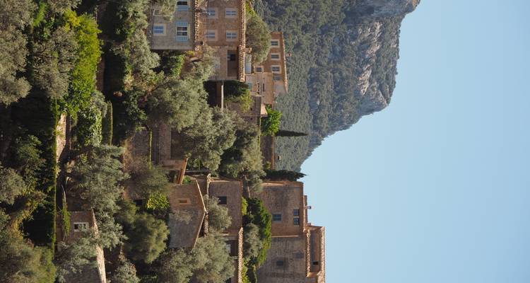 Des maisons en pierre en terrasses et des oliviers s'élevant sur une pente collinaire sous un arrière-plan de montagne rocheuse.