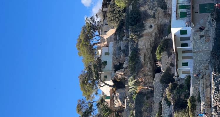 Groupe de maisons en falaise aux volets verts perchées sur des terrasses rocheuses sous un ciel bleu.
