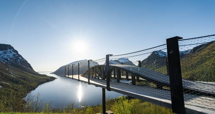 Passerelle avec point de vue architectural dominant un fjord ensoleillé avec des montagnes et un soleil éclatant.