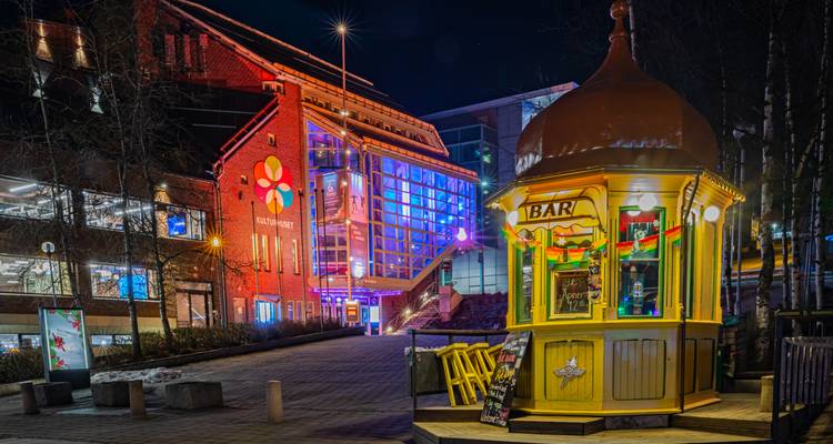 Scène de rue illuminée et colorée la nuit avec un petit kiosque de bar jaune et des bâtiments modernes.