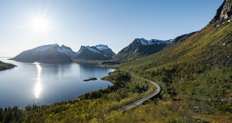 Vue aérienne élevée d'une route sinueuse, d'une forêt verte et d'un fjord réfléchissant avec un soleil bas et des pics.
