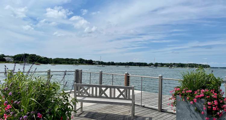 Une jetée en bois bordée de fleurs avec un banc offre une vue sur un port calme parsemé de voiliers.