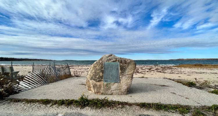 Un rocher commémoratif avec plaque se dresse sur une plage de sable exposée aux vents, au bord de la mer.