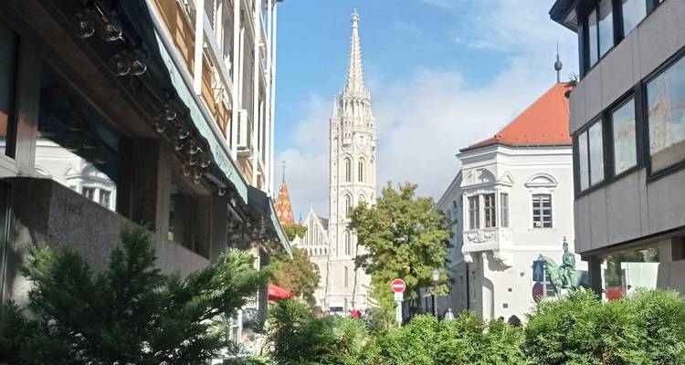 Matthias Church spire viewed down a sunny pedestrian street framed by green shrubs and historic buildings.