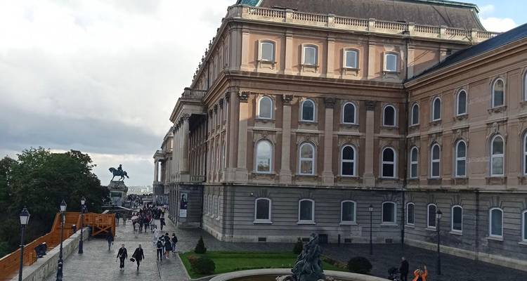 Visitors walking between the wings of Buda Castle near an ornate fountain on a cloudy afternoon.