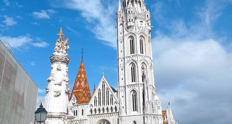 Tall white Holy Trinity Column and Matthias Church tower rising into a bright blue sky with scattered clouds.