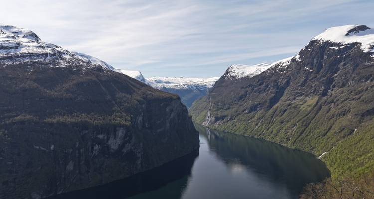 Vue classique d'un fjord étroit et ombragé bordé de falaises parsemées de neige sous un ciel doux.