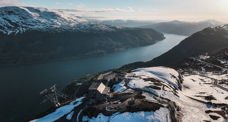 Station au sommet enneigée du Loen Skylift dominant un fjord bleu s'étendant vers les chaînes de montagnes lointaines et brumeuses au coucher du soleil.
