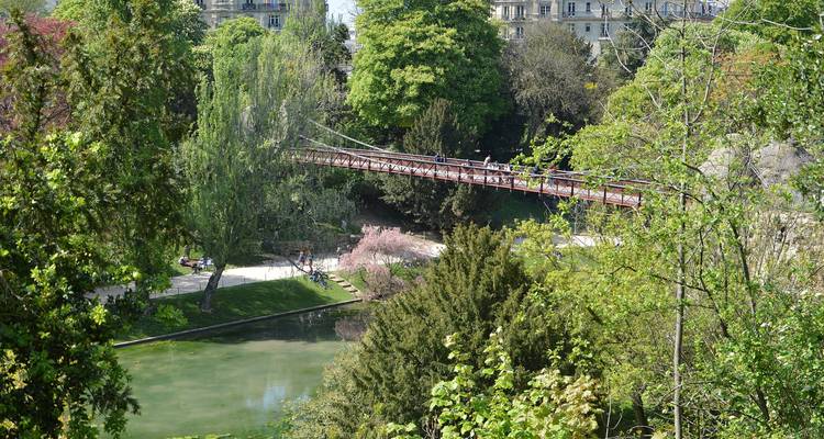 Scène luxuriante d'un parc parisien avec un pont piétonnier rouge traversant un ravin verdoyant et un étang entourés d'une végétation dense.