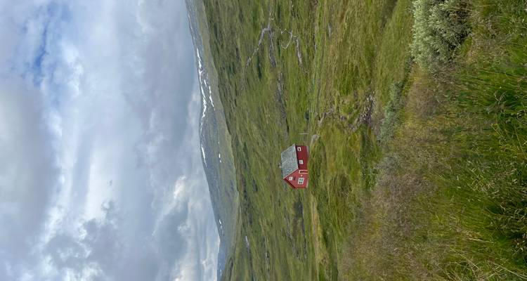 Une petite cabane rouge isolée se dresse au milieu de vastes prairies verdoyantes des Highlands sous un ciel nuageux avec des plaques de neige sur les collines lointaines.