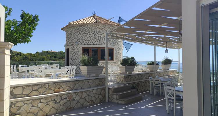 Sunlit stone terrace with white tables and chairs overlooking the sea from a circular building.