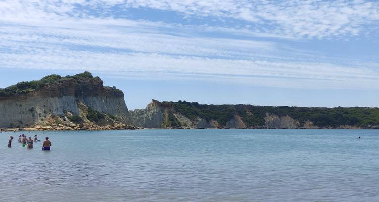 Clear blue bay flanked by white cliffs with swimmers enjoying shallow water under light clouds.
