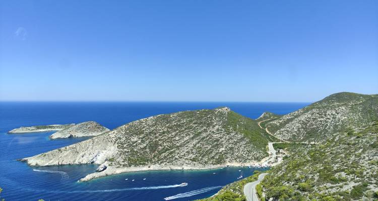 Panoramic coastal vista of steep green hills, winding road, and deep blue Ionian Sea.