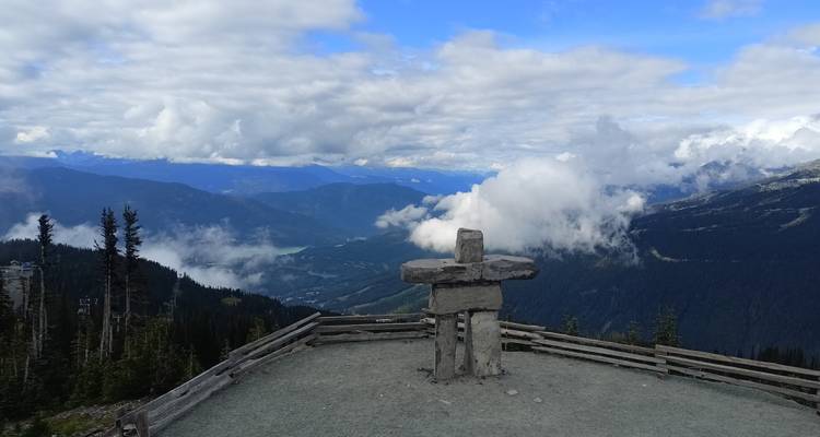 Stone Inukshuk statue at mountain viewpoint overlooking deep valleys and distant peaks.
