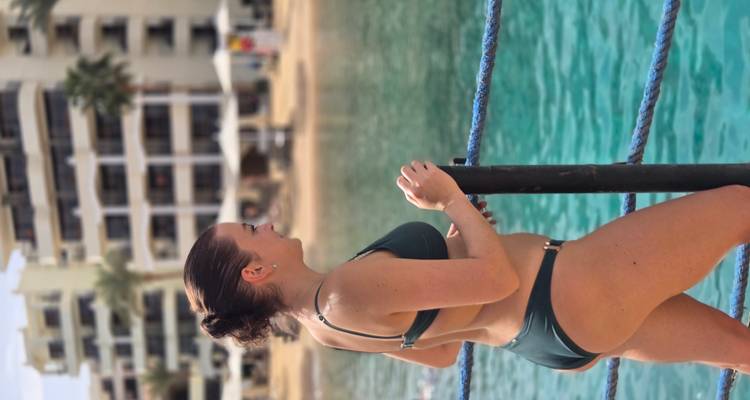 Woman in swimwear standing by a rope railing, gazing over clear lagoon toward a resort.