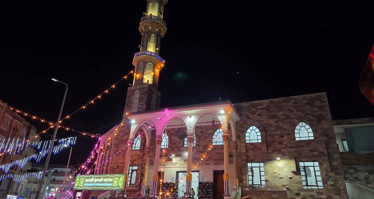Stone-clad mosque with tall minaret lit up at night, festive lights strung across the street.