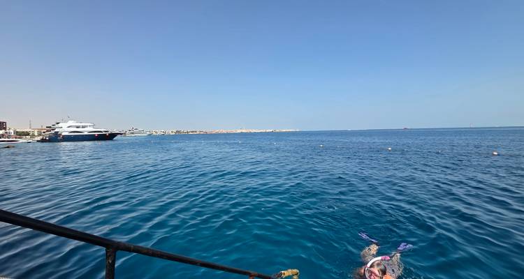 Wide seascape with snorkeler near ladder and yachts anchored in the distance on a clear day.