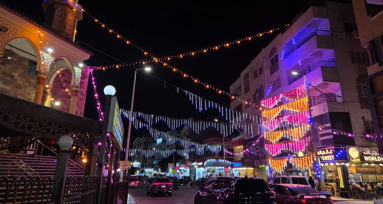 Busy city street at night with colorful string lights and passing traffic beside a mosque.