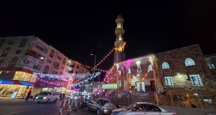 Nighttime cityscape featuring a tall minaret, festive lights, cars and busy pedestrians.