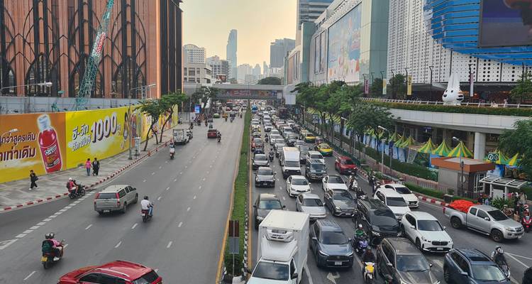 Avenida concurrida de Bangkok llena de autos y motos, rodeada de edificios altos y vallas publicitarias