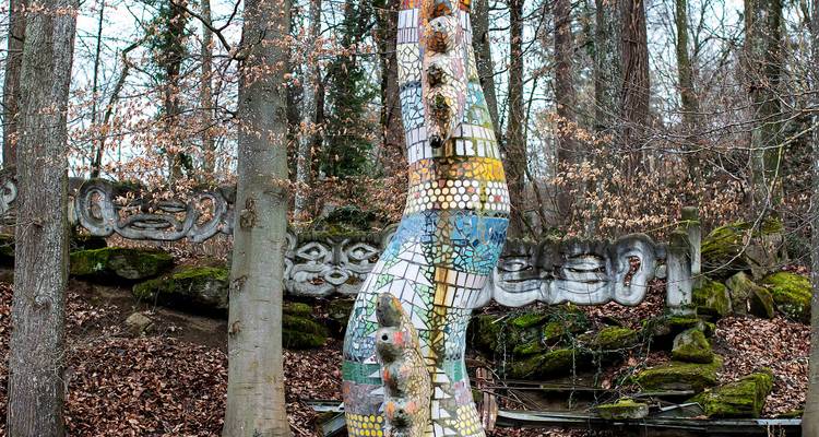 Colorful mosaic serpent sculpture winding between bare trees and mossy stones in a forest park.